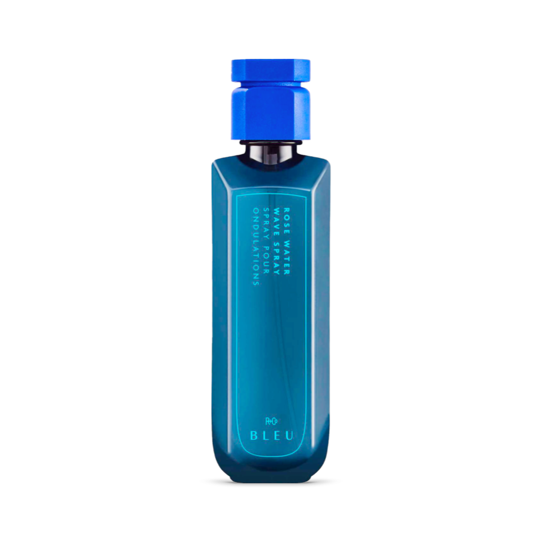 Elegant navy blue bottle of R+Co BLEU Rose Water Wave Spray, 6.8oz, with a bright blue cap. The front label is adorned with the product name and details in reflective silver and teal fonts, showcasing a sleek and high-end design.